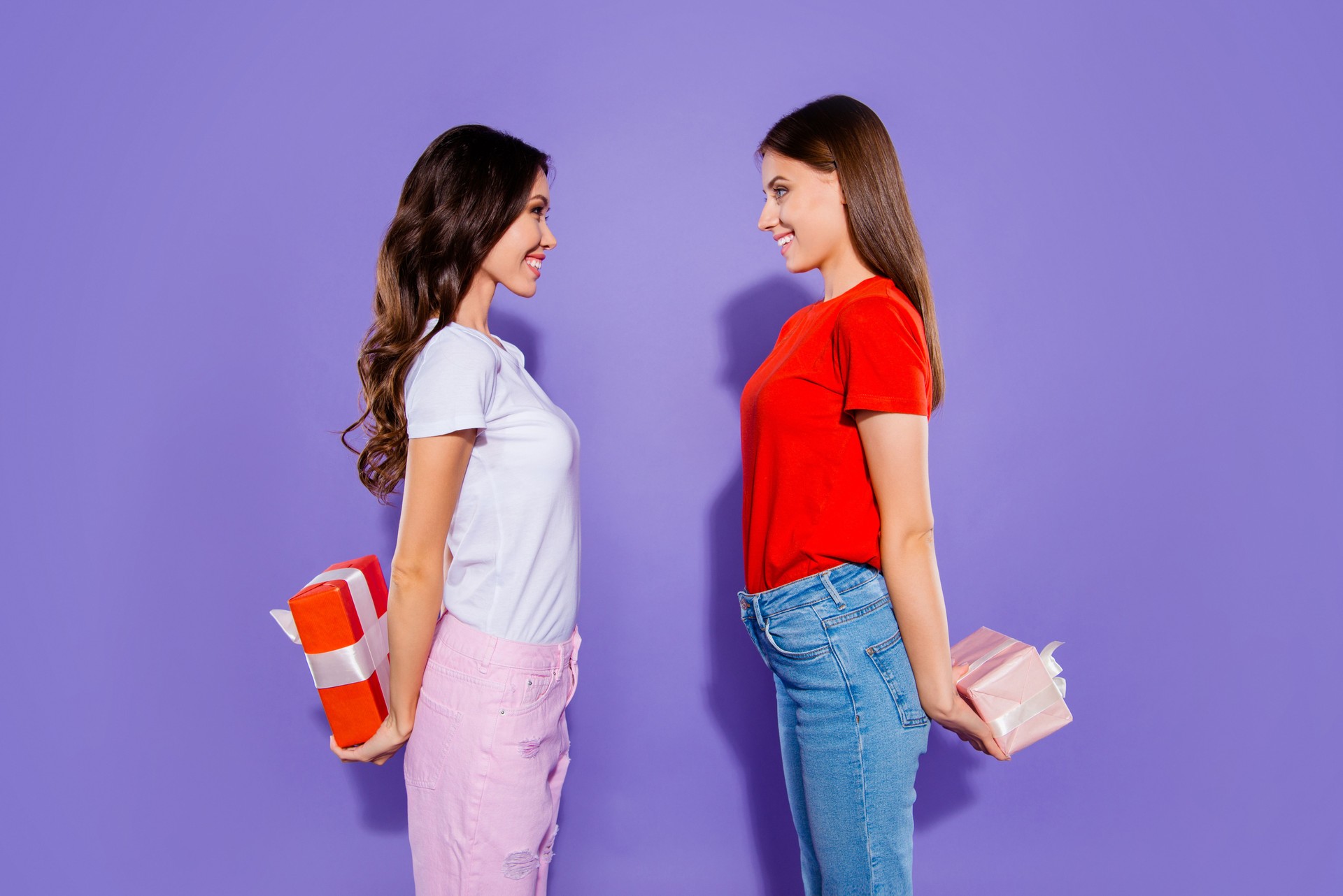 Profile side view portrait of two nice dreamy adorable sweet attractive cheerful girls holding in hands hiding behind back bow ribbon boxes having fun isolated over violet purple pastel background