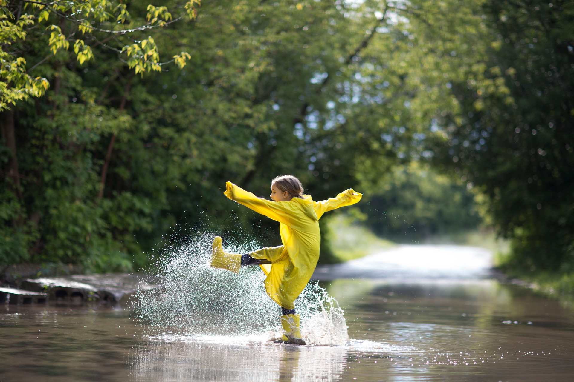 Happy An eight-year-old girl wearing yellow rubber boots and a yellow raincoat Jumping in a puddle after a summer rain